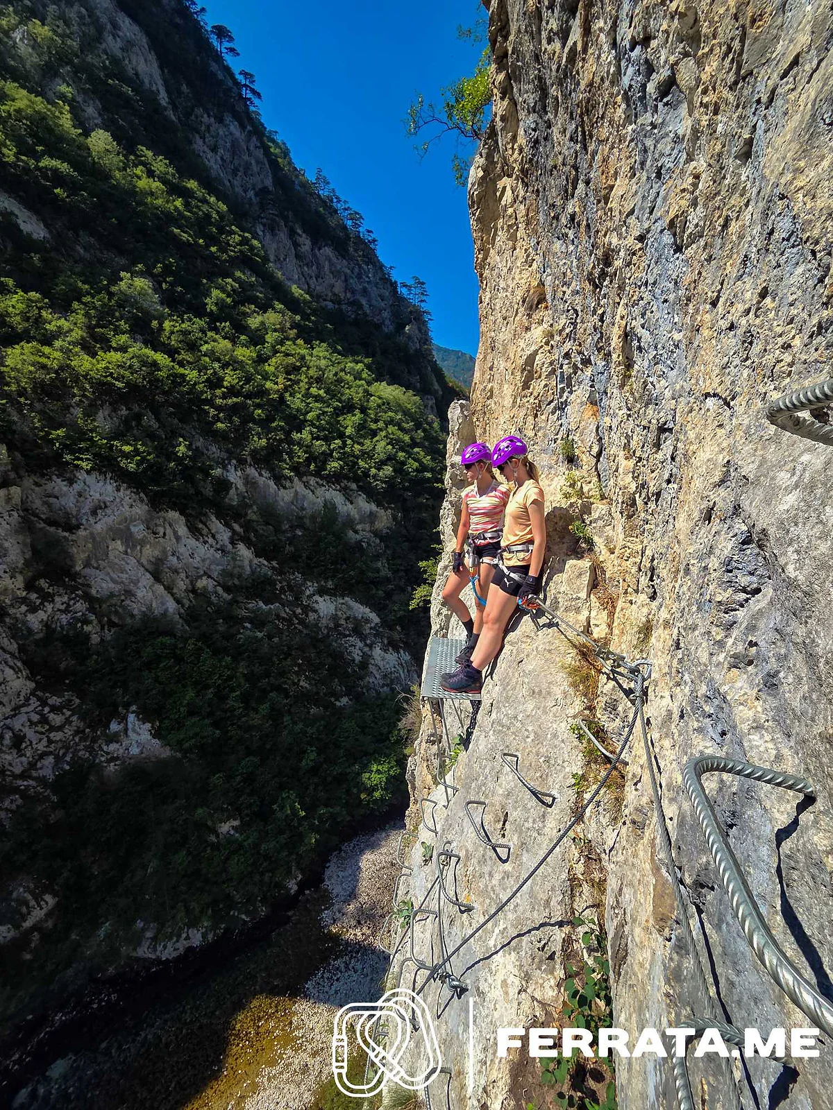 Two climbers in purple helmets on the steel bench halfway up the wall, with the canyon dropping away below them.