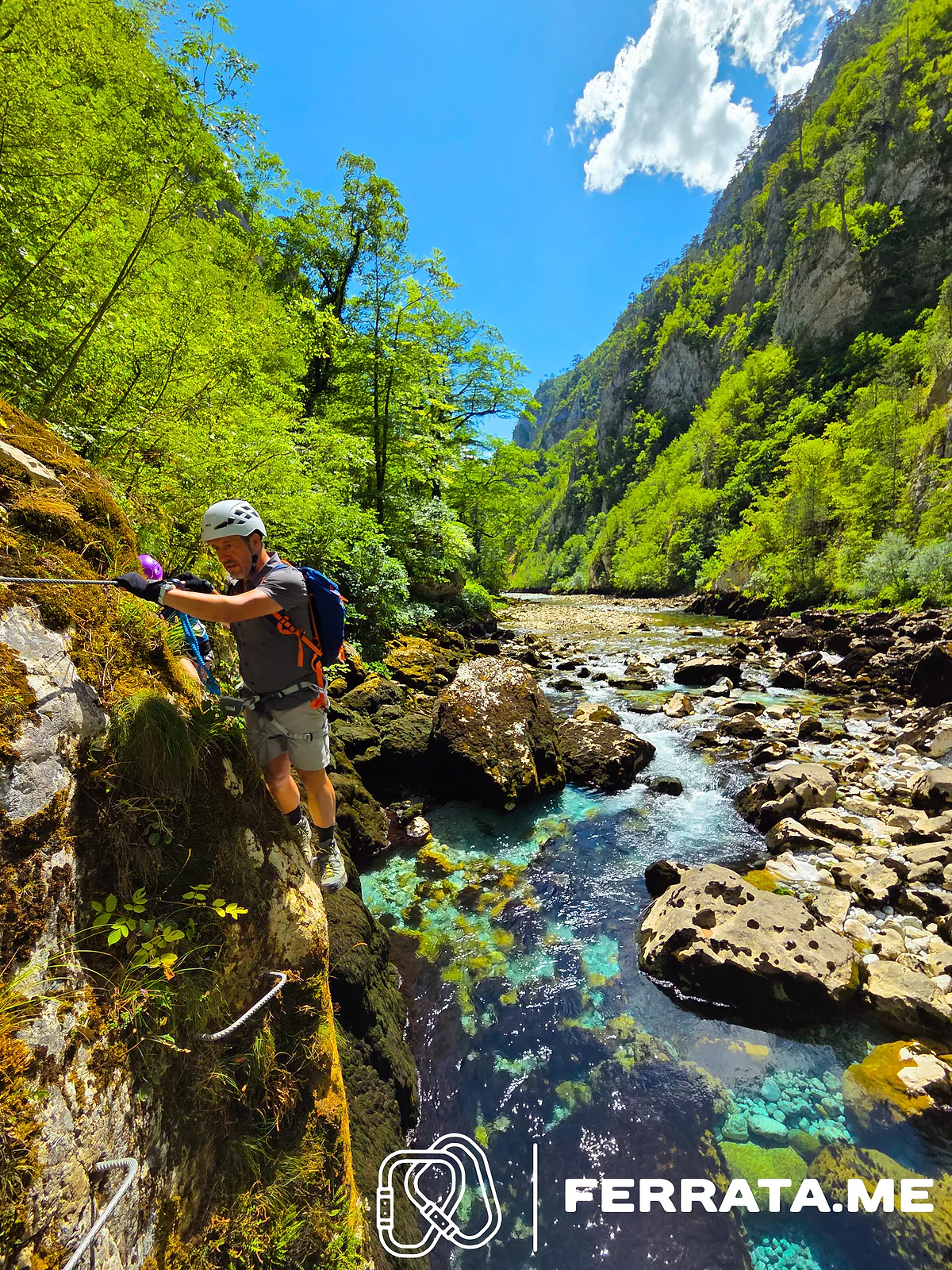 A climber in a white helmet and blue backpack holds the cable on a mossy rock wall above clear turquoise pools of the Piva, with a second climber behind him and the green canyon beyond.