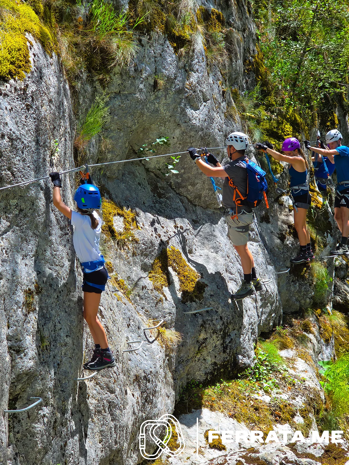 A girl in a blue helmet and adults in white and purple helmets move sideways along a horizontal cable on pale grey rock, standing on iron pegs.