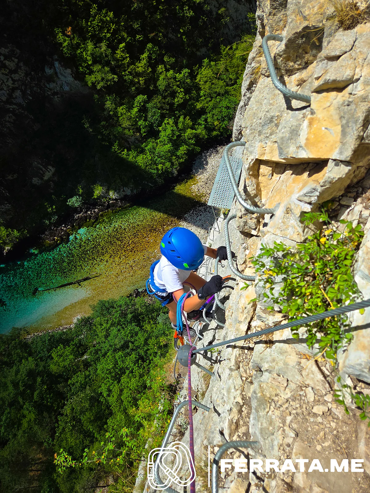 A young climber in a blue helmet on iron rungs above an emerald pool.