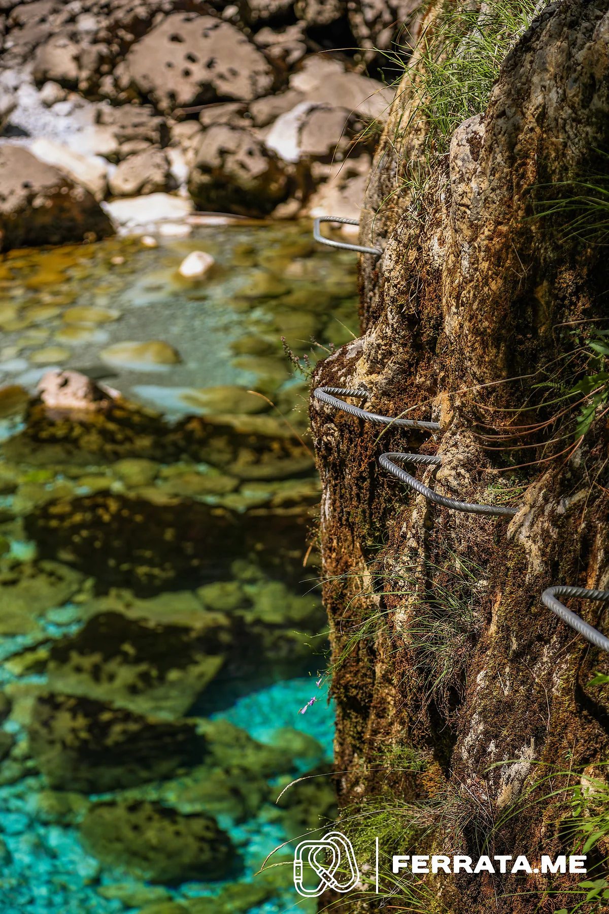 Bent steel staples set into a mossy rock face beside a shallow, clear pool with pale stones on the bottom.