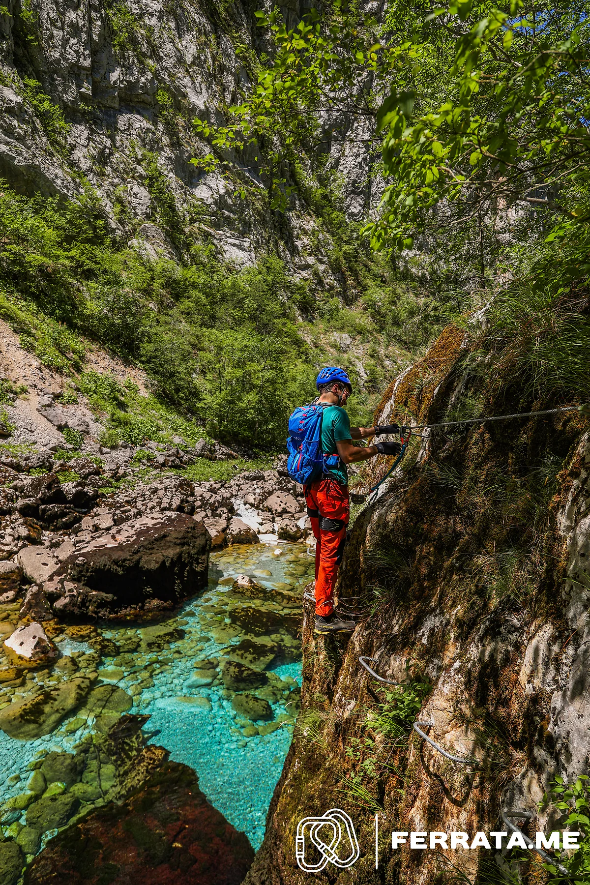 A man in a blue helmet, green top and red trousers, with a blue backpack, holds the cable as he steps along the wall above a clear turquoise pool.
