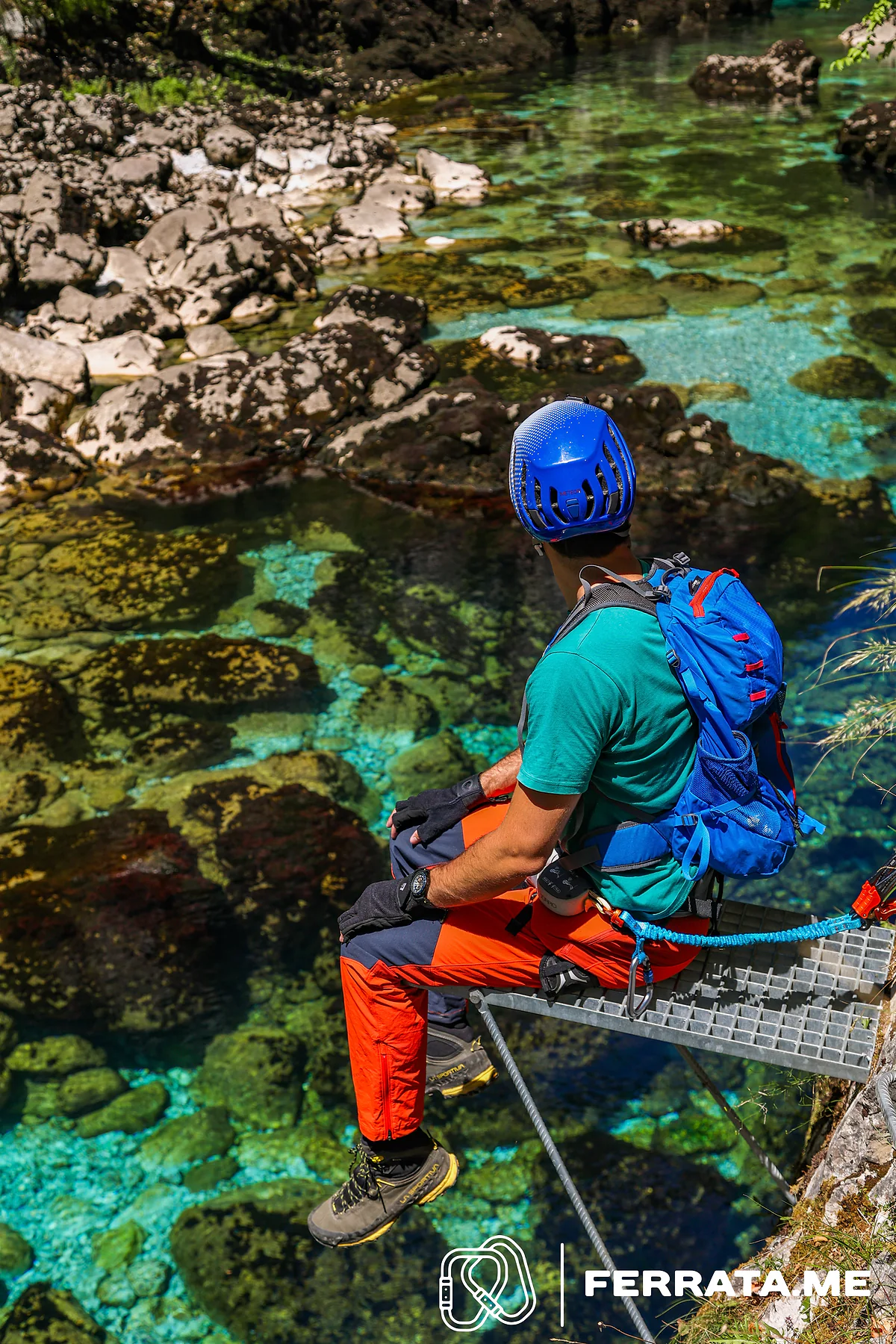 A climber sitting on a steel grate above turquoise water, clipped to the cable.