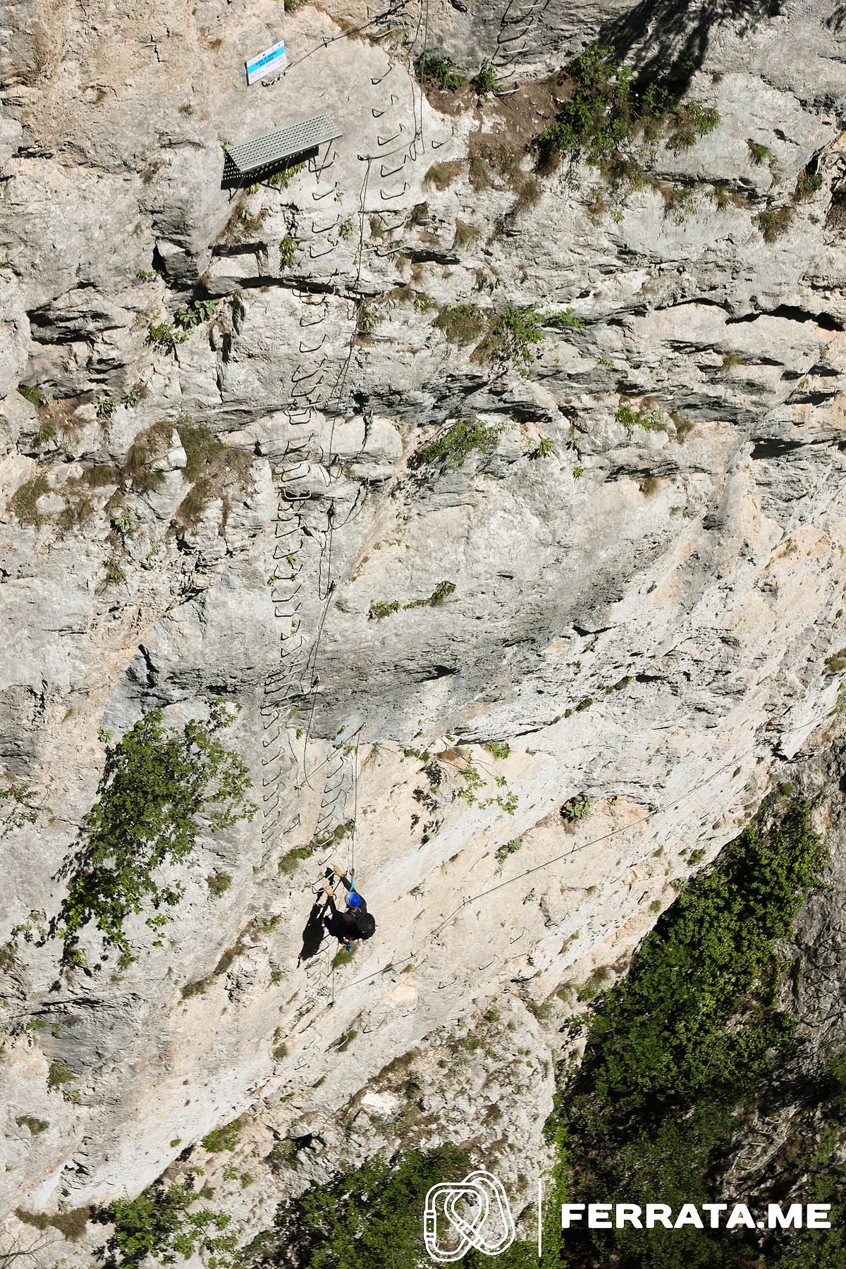 Seen from above, a single climber in a blue helmet climbs the iron rungs on a wide pale limestone face, with the steel bench and its sign higher up the wall.