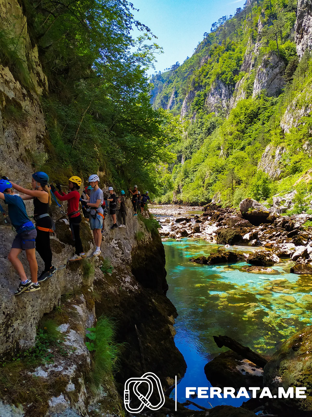 A line of climbers in blue, yellow and white helmets clipped to the cable along a mossy rock wall above a clear turquoise river, with the forested canyon stretching away ahead.