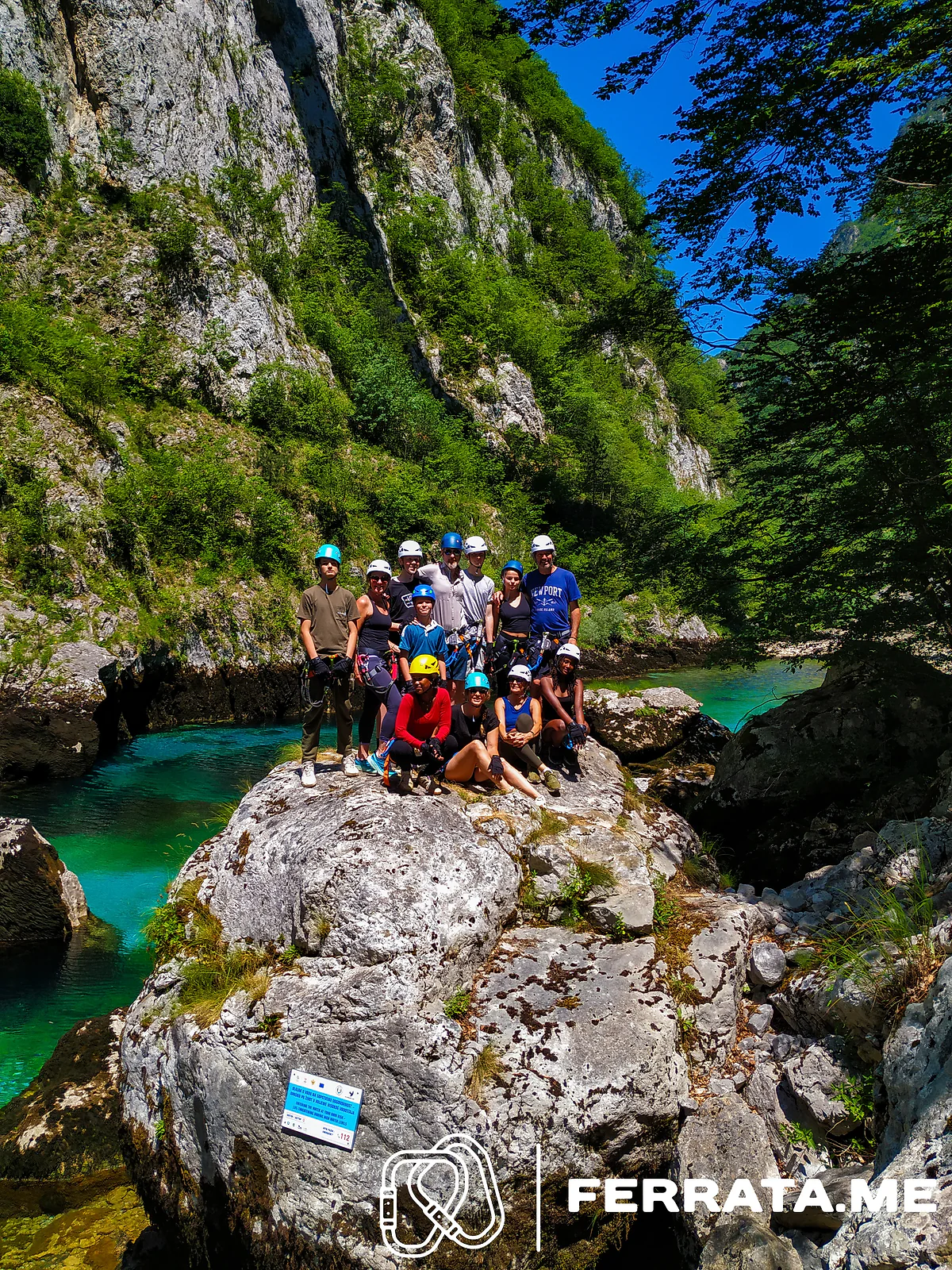 A big group in helmets on a boulder above turquoise water.