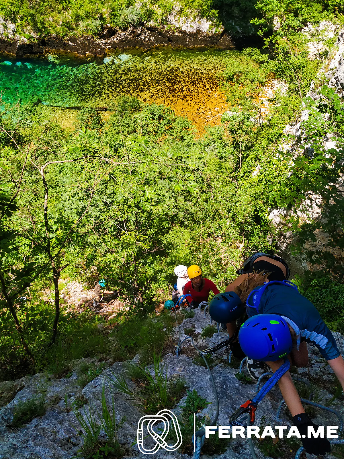 Climbers in blue helmets at the start of the wall, seen from above, with trees and the river below.