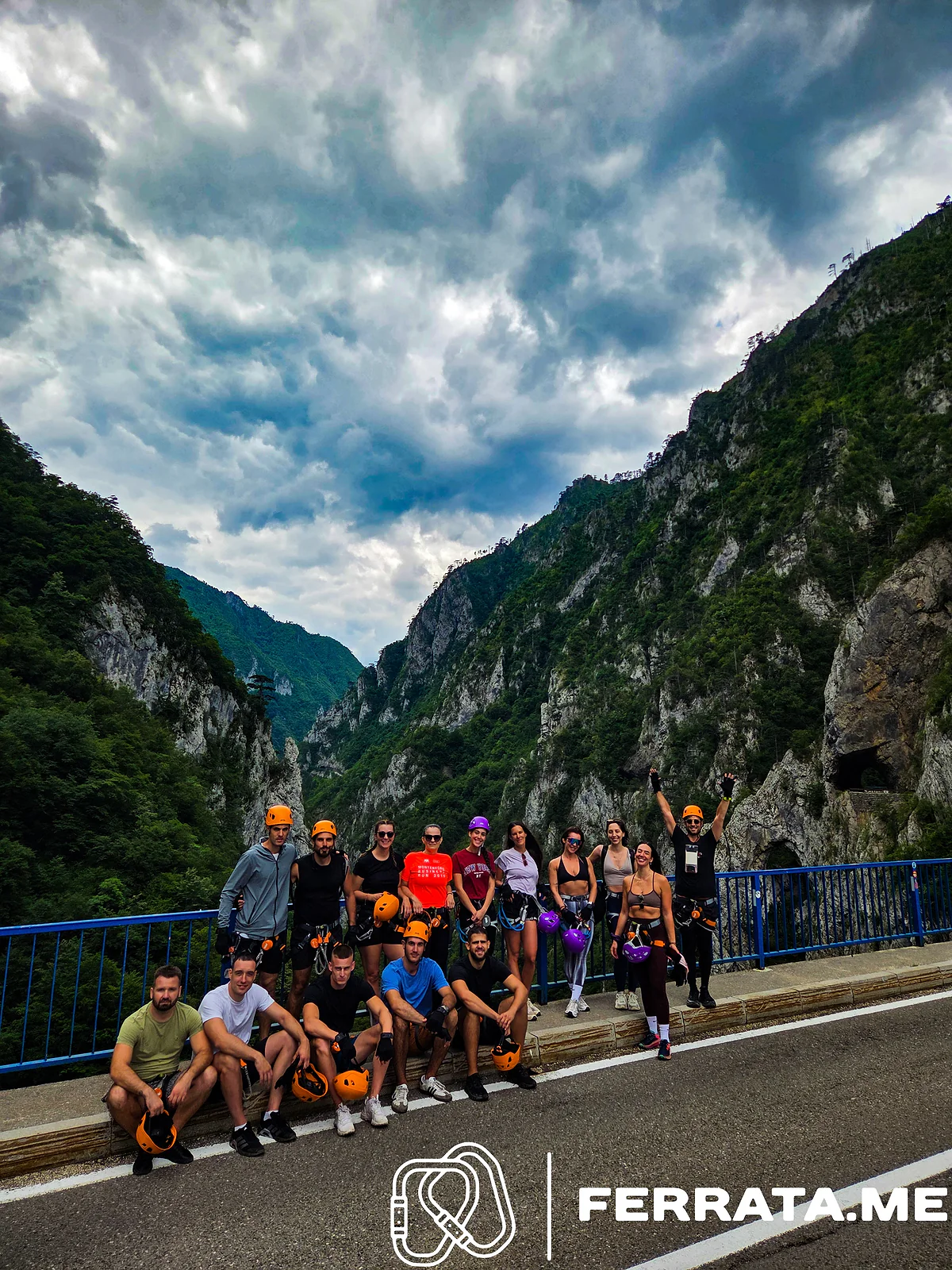 About twenty people in climbing harnesses, most holding orange or purple helmets, pose by the blue railings of the bridge, with the green walls of Piva Canyon rising behind them under a cloudy sky.