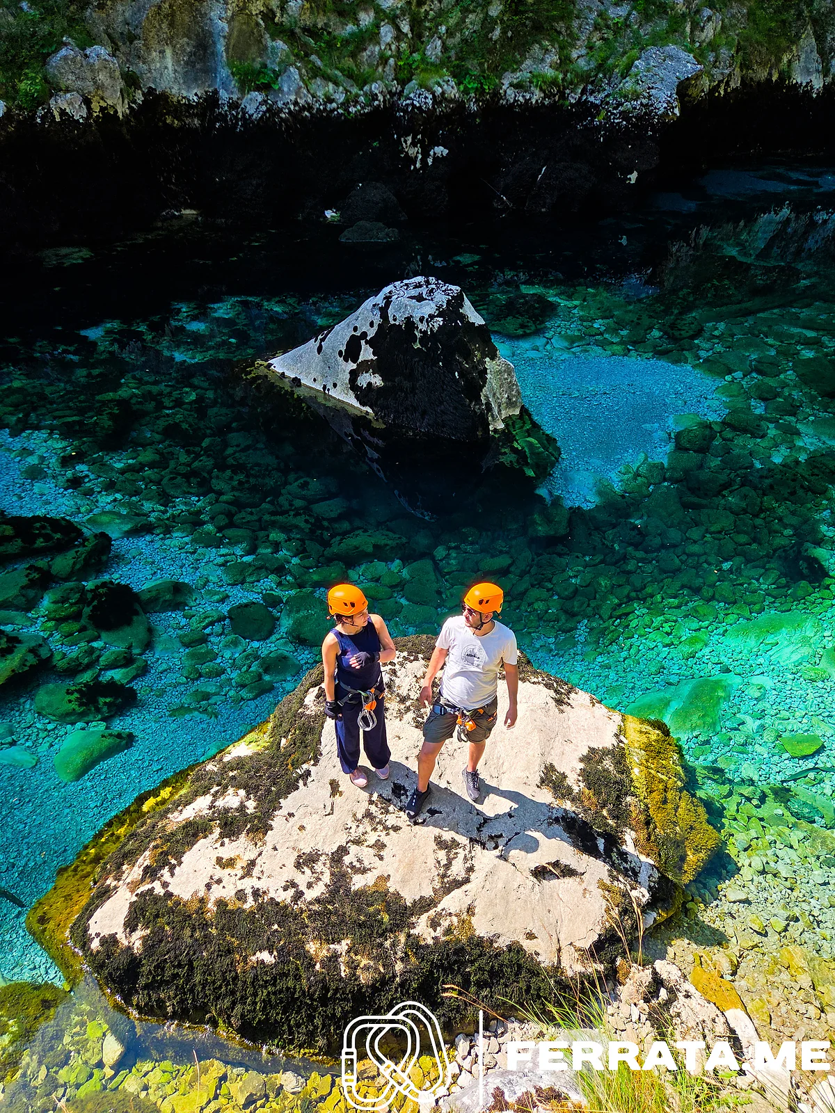 Seen from above, a woman and a man in orange helmets and climbing harnesses stand on a pale boulder in a clear turquoise pool of the Piva, with stones visible on the riverbed.