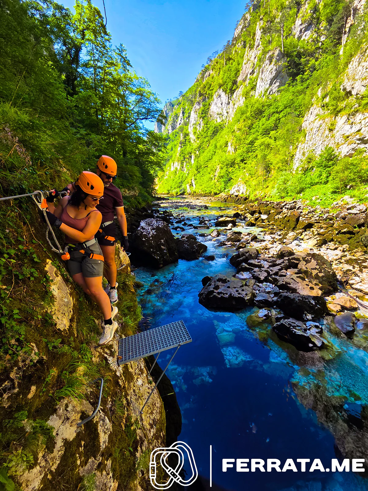 A woman and a man in orange helmets clipped to the cable on the canyon wall look down at a steel grate over a deep blue pool, with the rocky river and green canyon walls beyond.