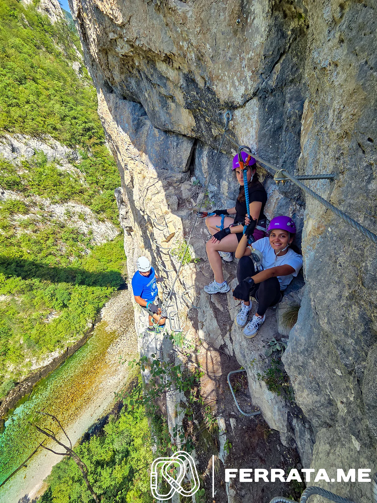 Two women in purple helmets rest in a recess of the wall, clipped to the cable, while a climber in a white helmet comes up the rungs below them and the river runs far below on the left.