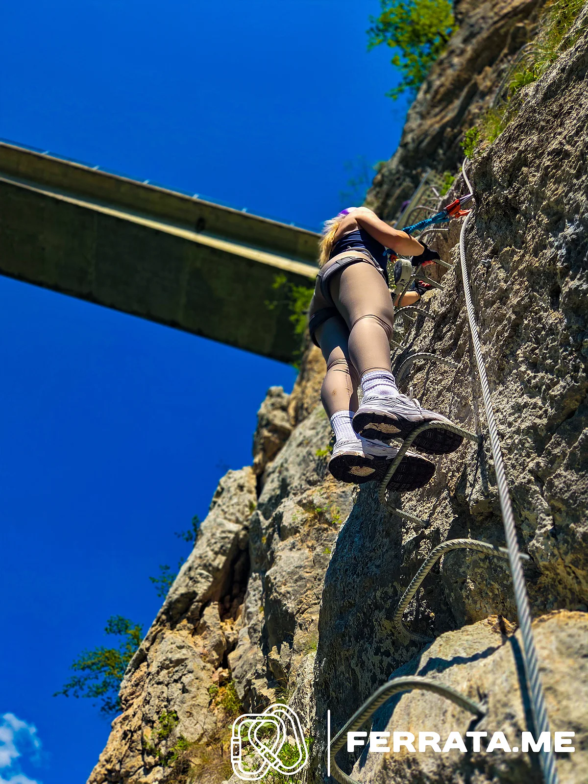 Looking up from below at a climber in grey leggings and white trainers on iron rungs beside the steel cable, with the concrete bridge overhead against a deep blue sky.