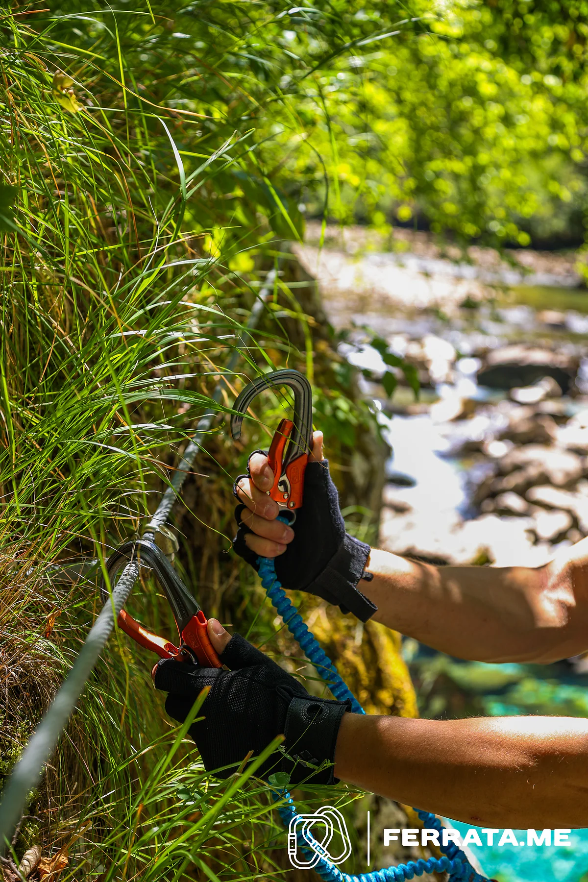 Two gloved hands on the cable: one squeezes the orange lever of a carabiner still clipped to the cable, the other holds the second carabiner open, with the blue elastic arms of the set below and the river in the background.