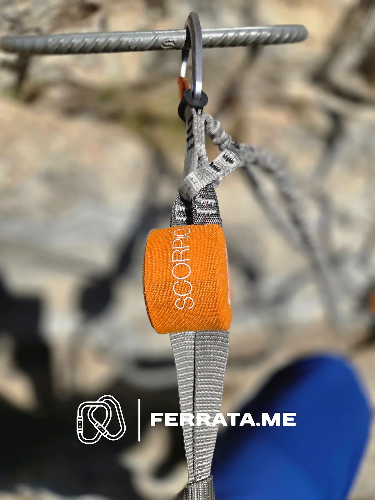 An orange Petzl Scorpio energy absorber hangs from a grey carabiner clipped to an iron rung, with the grey webbing of the lanyard above and below it.