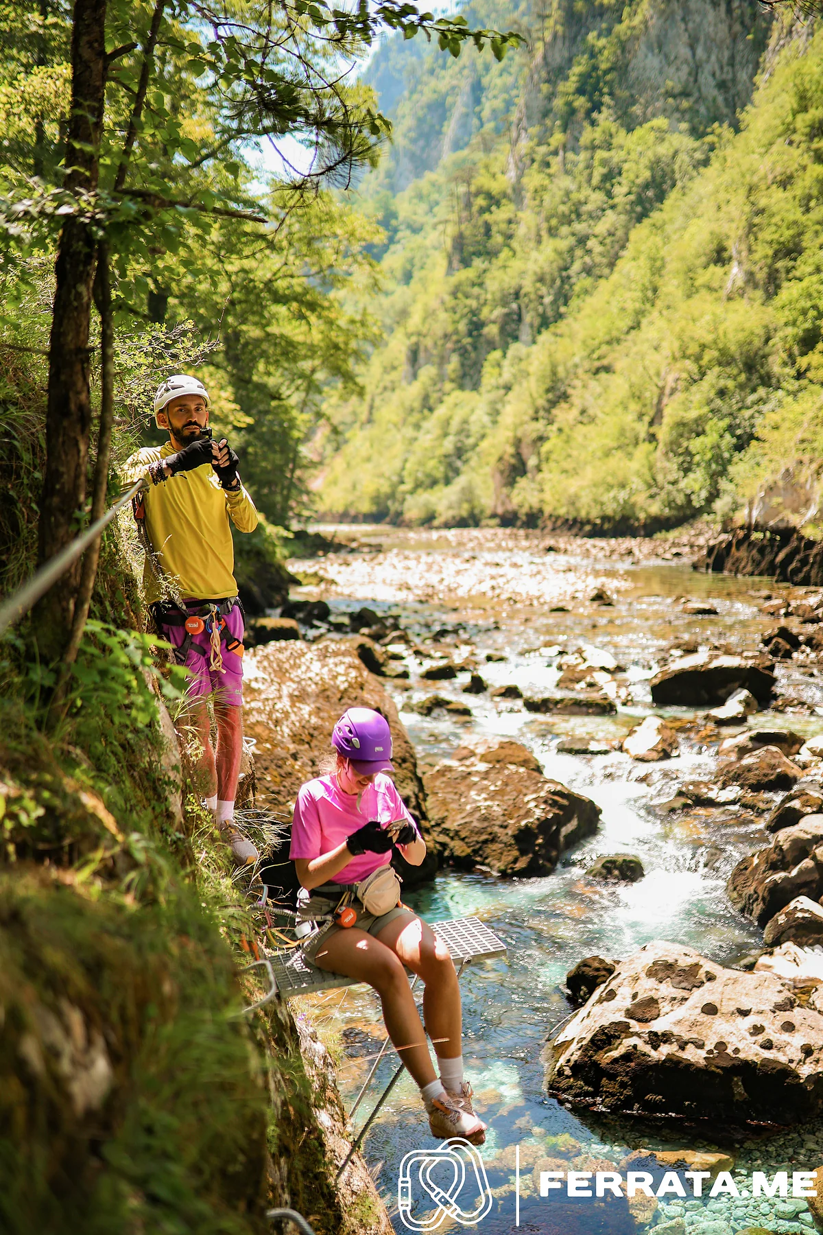 A woman in a purple helmet and pink T-shirt sits on a steel grate with her feet over the shallow river, while a man in a white helmet and yellow top stands on the rock behind her, holding up a phone.