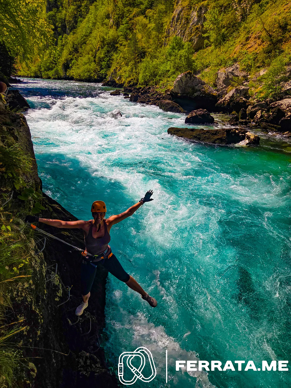 A woman in an orange helmet clipped to the cable on the canyon wall throws her arms out over fast white water and turquoise pools of the Piva, with green slopes behind.