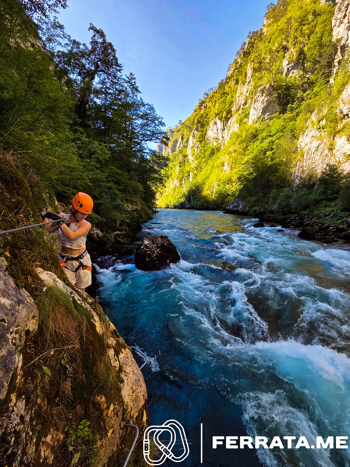 A woman in an orange helmet clipped to the cable on a ledge above fast blue rapids, with green trees and sunlit limestone walls up the canyon under a clear sky.