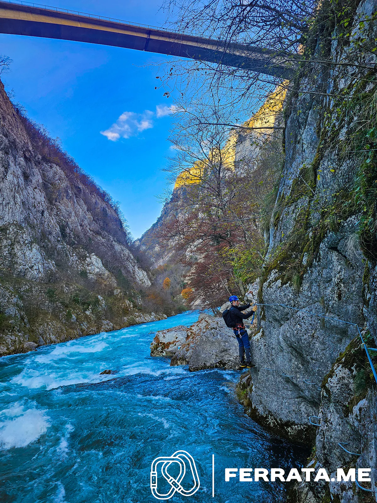 A climber in a blue helmet, a dark jacket and long blue trousers, with a small backpack, holds the cable on a rock beside fast blue rapids, with bare trees and the bridge high above.