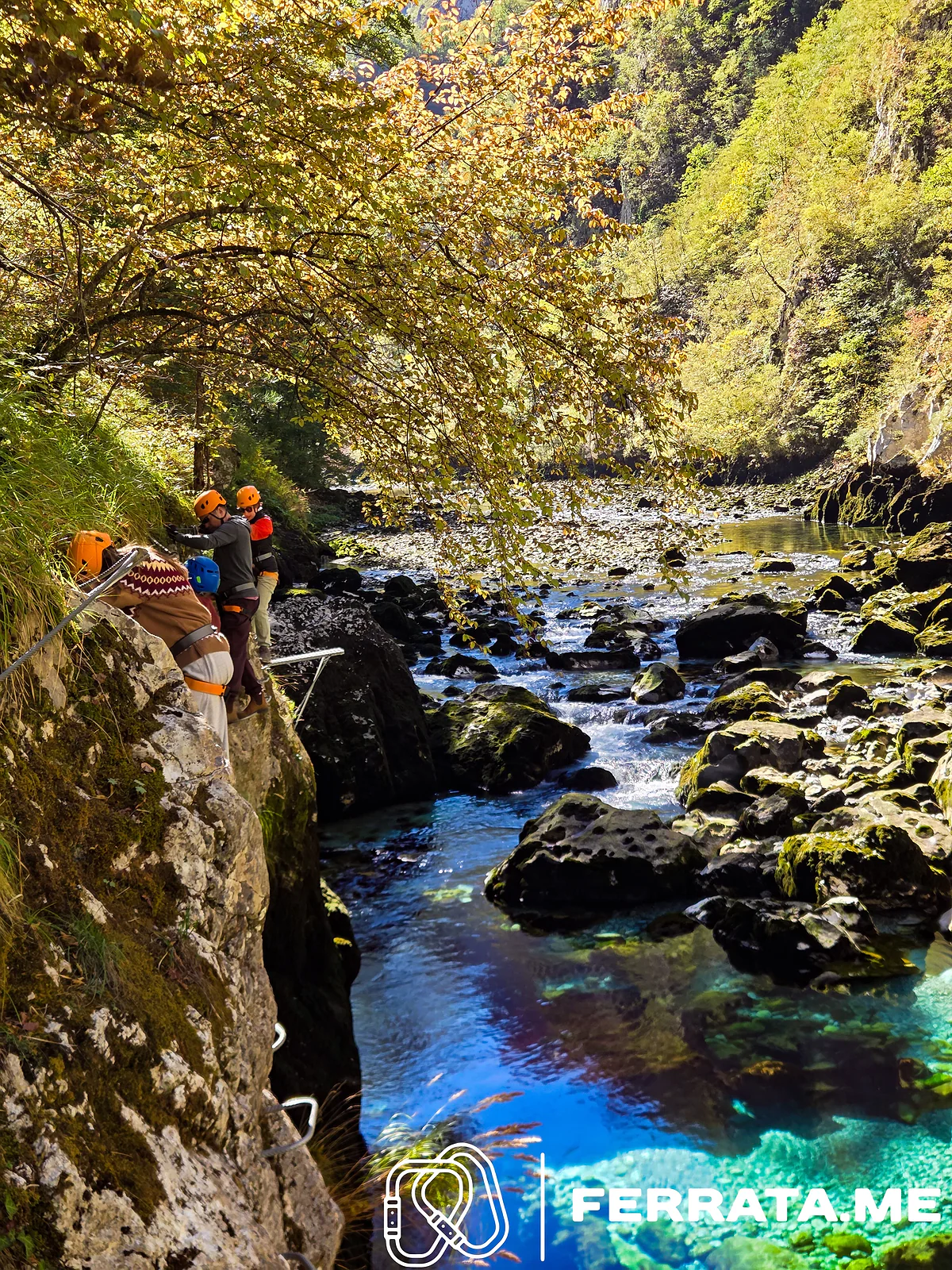 Climbers in orange and blue helmets clipped to the cable on a mossy rock ledge under yellowing autumn leaves, above clear blue pools and boulders in the river.