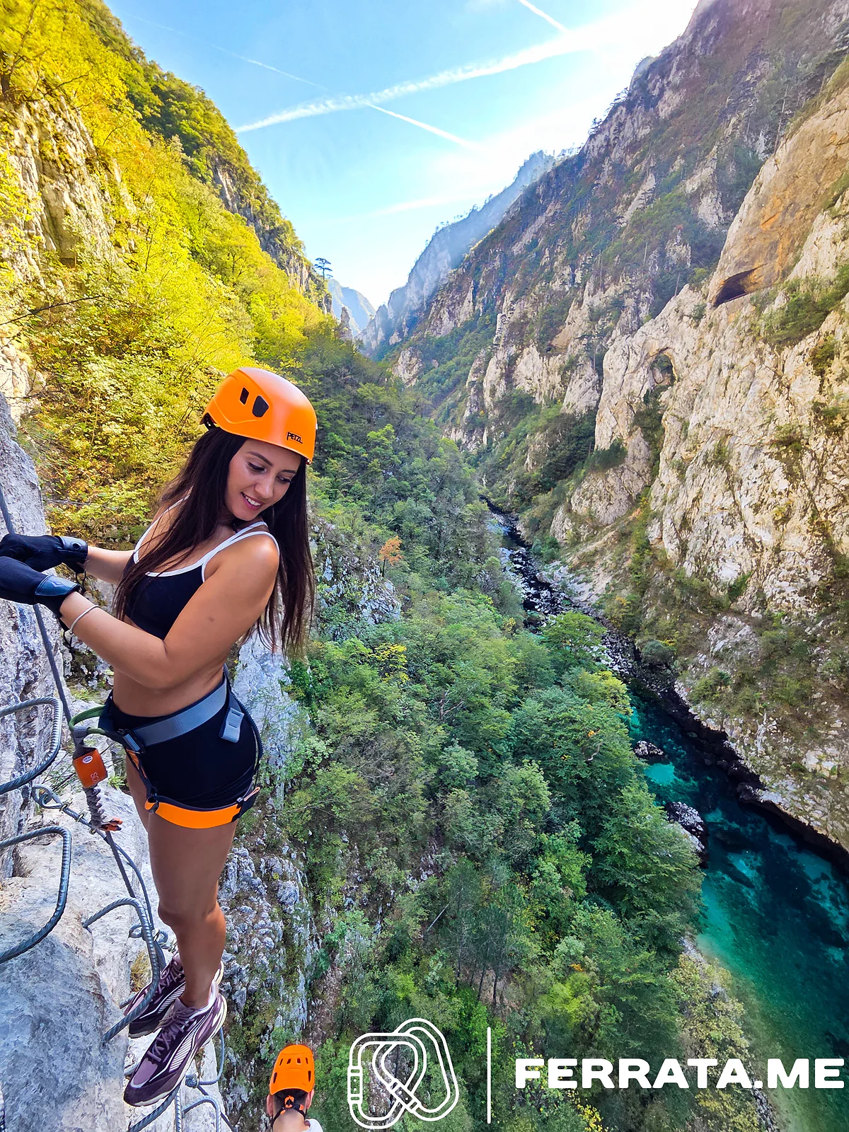 A woman in an orange helmet and black top stands on iron rungs high on a limestone wall, with the green Piva canyon and a turquoise river stretching away below.