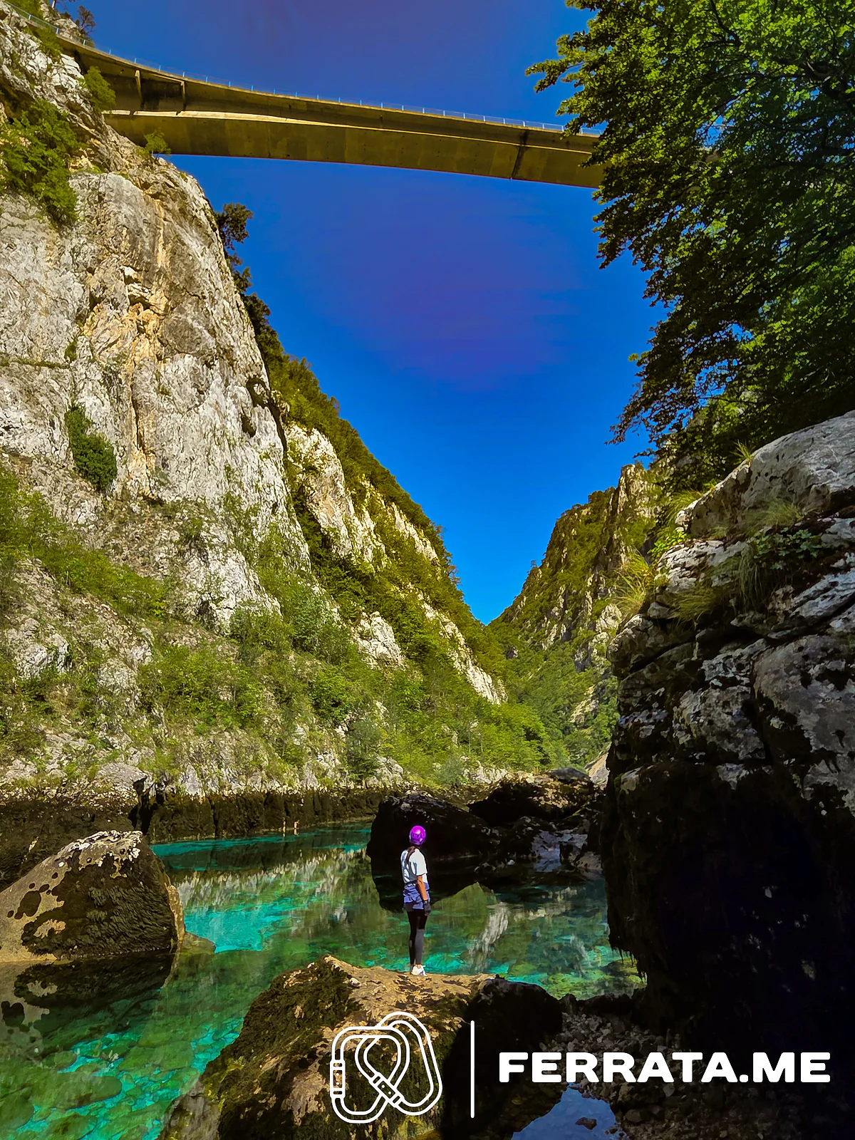 A woman in a purple helmet stands on a rock beside a clear turquoise pool at the bottom of the canyon, with the road bridge high overhead and grey limestone walls on both sides.