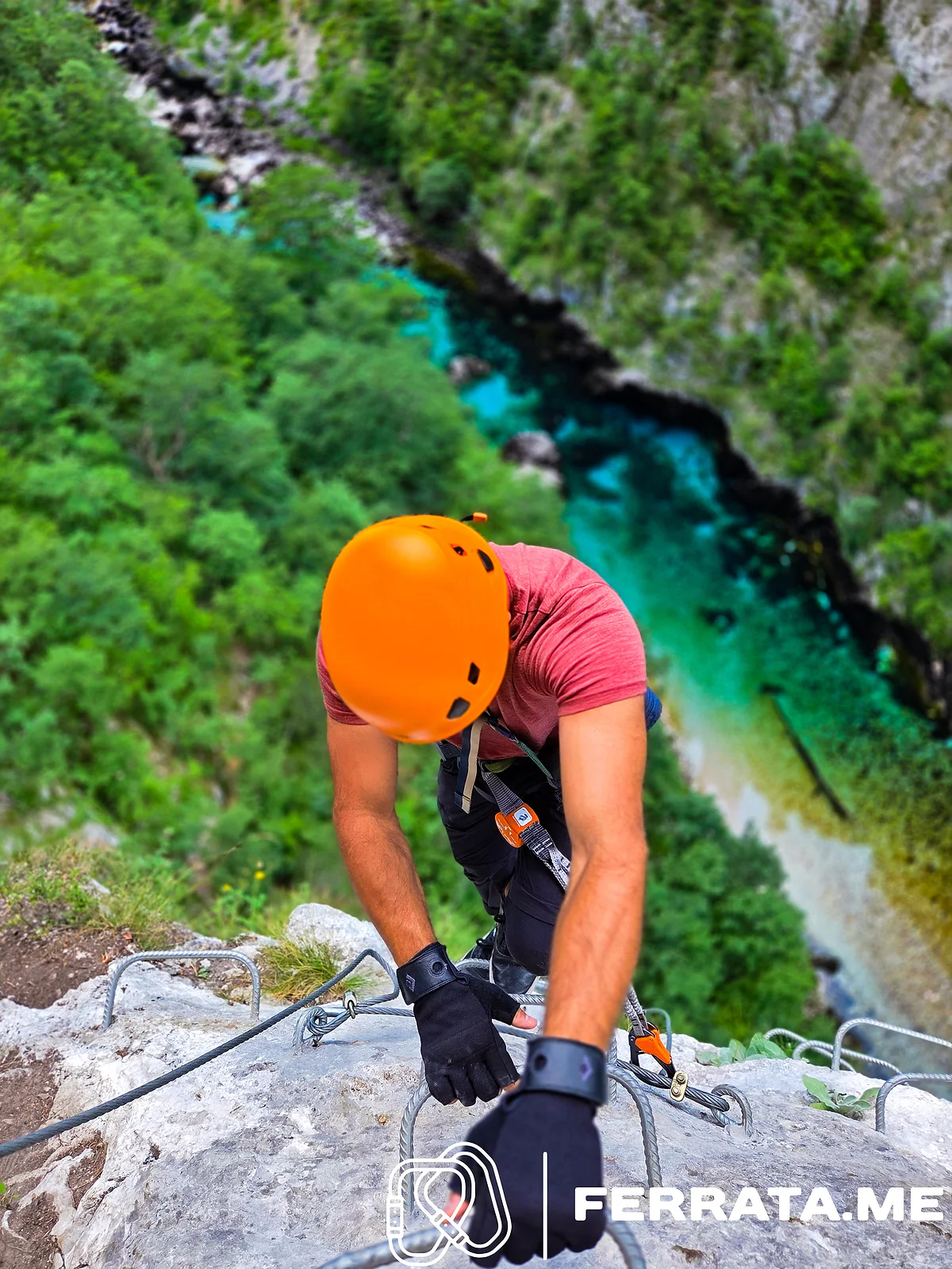 Looking straight down at a man in an orange helmet and a faded red T-shirt pulling himself up on iron rungs, with the turquoise river far below between forested slopes.