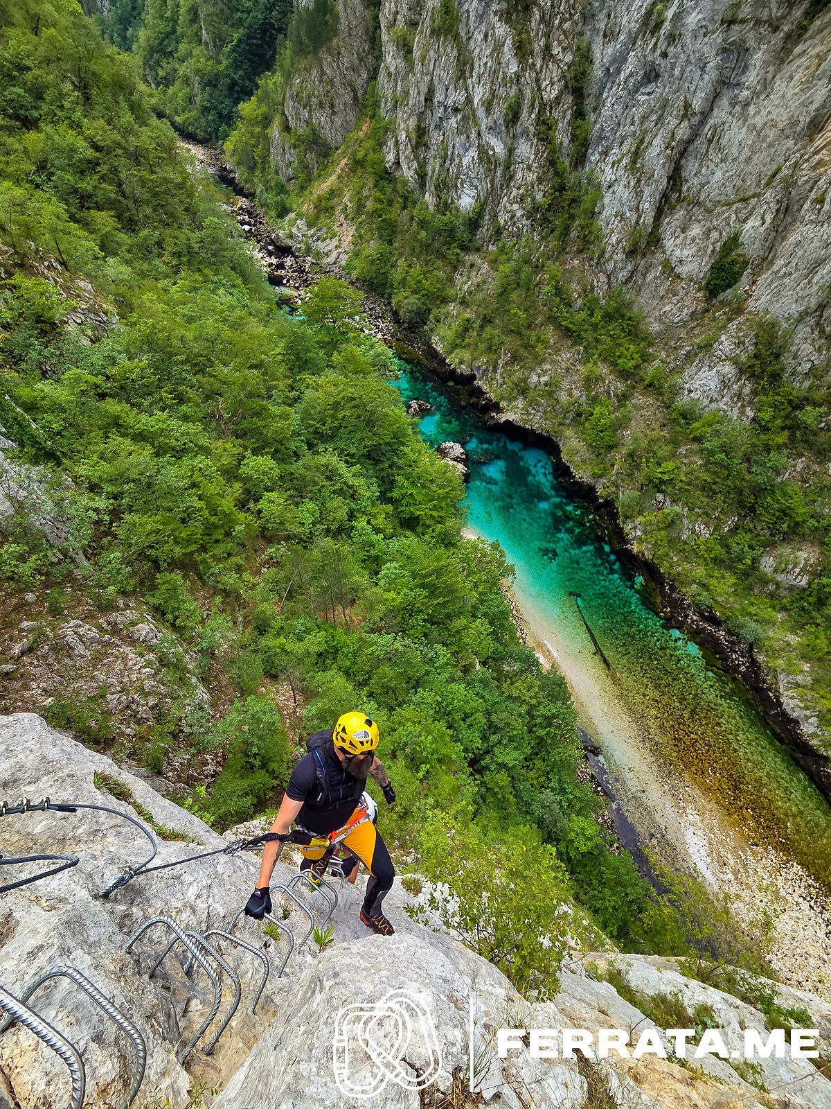 A climber in a yellow helmet and black top steps up iron rungs on pale rock, with the turquoise river far below running down a deep green canyon between grey cliffs.