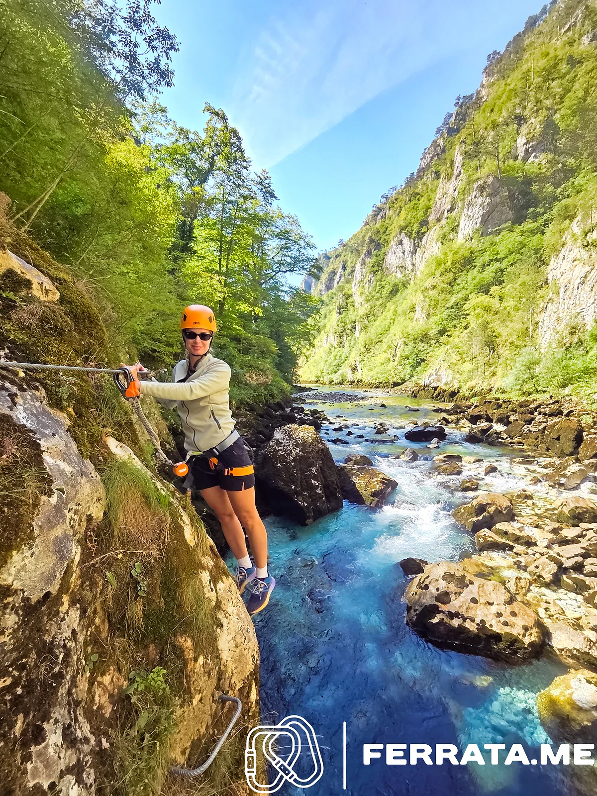 A woman in an orange helmet, sunglasses, a light zip-up jacket and black shorts holds the cable on the traverse, with fast clear water below her and green canyon walls under a blue sky.