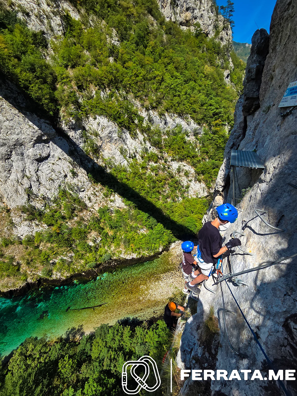 Two young climbers in blue helmets climb iron rungs on the wall above a climber in an orange helmet, with the green river and the grey canyon side far below.