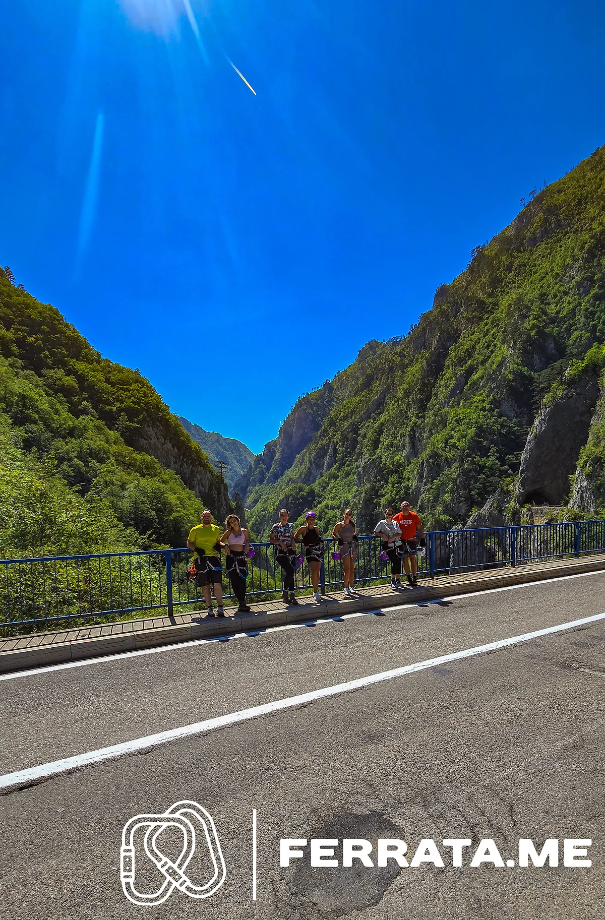 Seven people in climbing harnesses stand at the blue railing of the road bridge, with the forested walls of Piva Canyon rising on both sides under a clear blue sky.