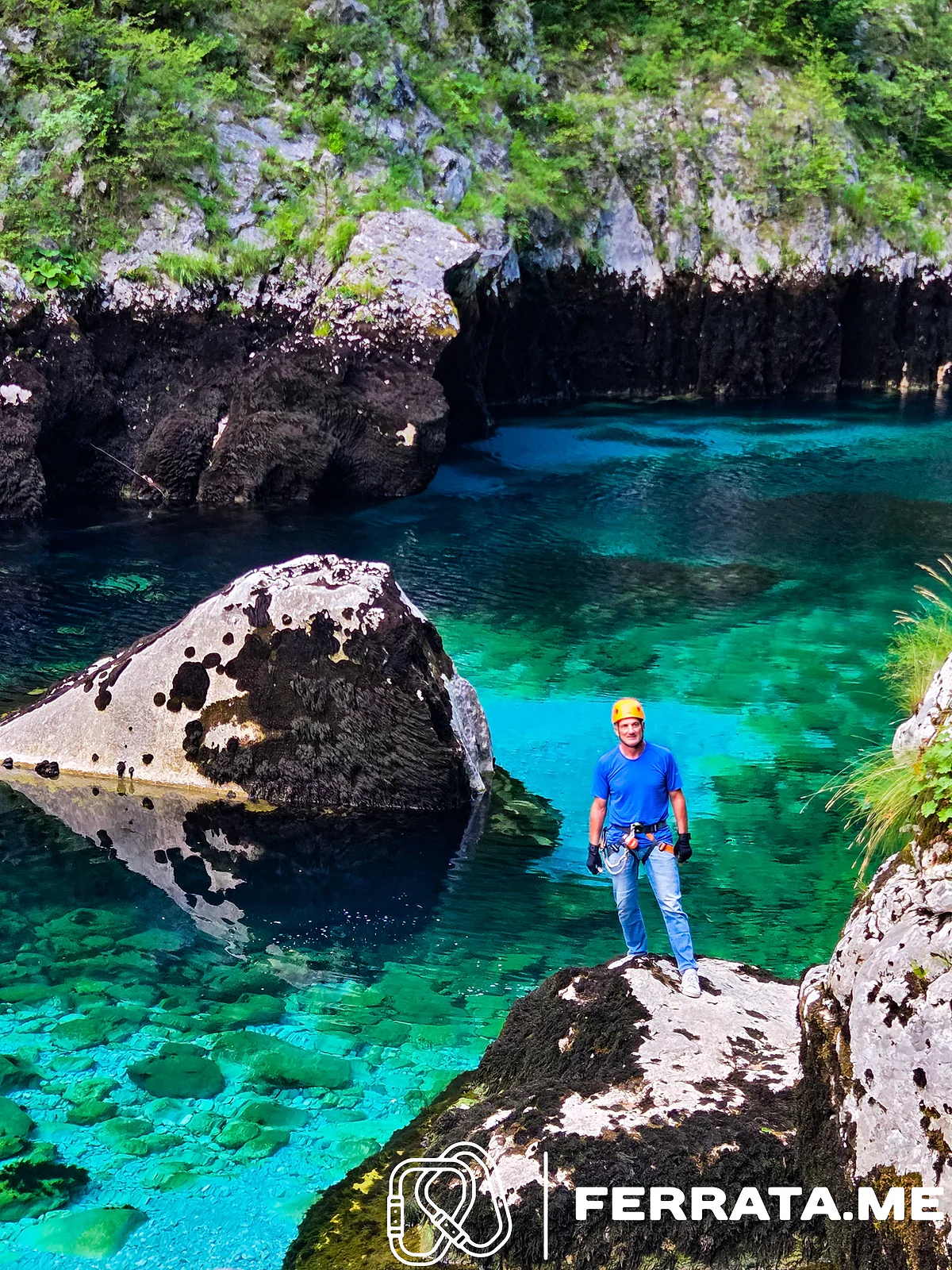 A man in a yellow helmet, blue T-shirt and climbing harness stands on a mossy boulder at the edge of a clear turquoise pool, with a large black and white boulder in the water beside him.