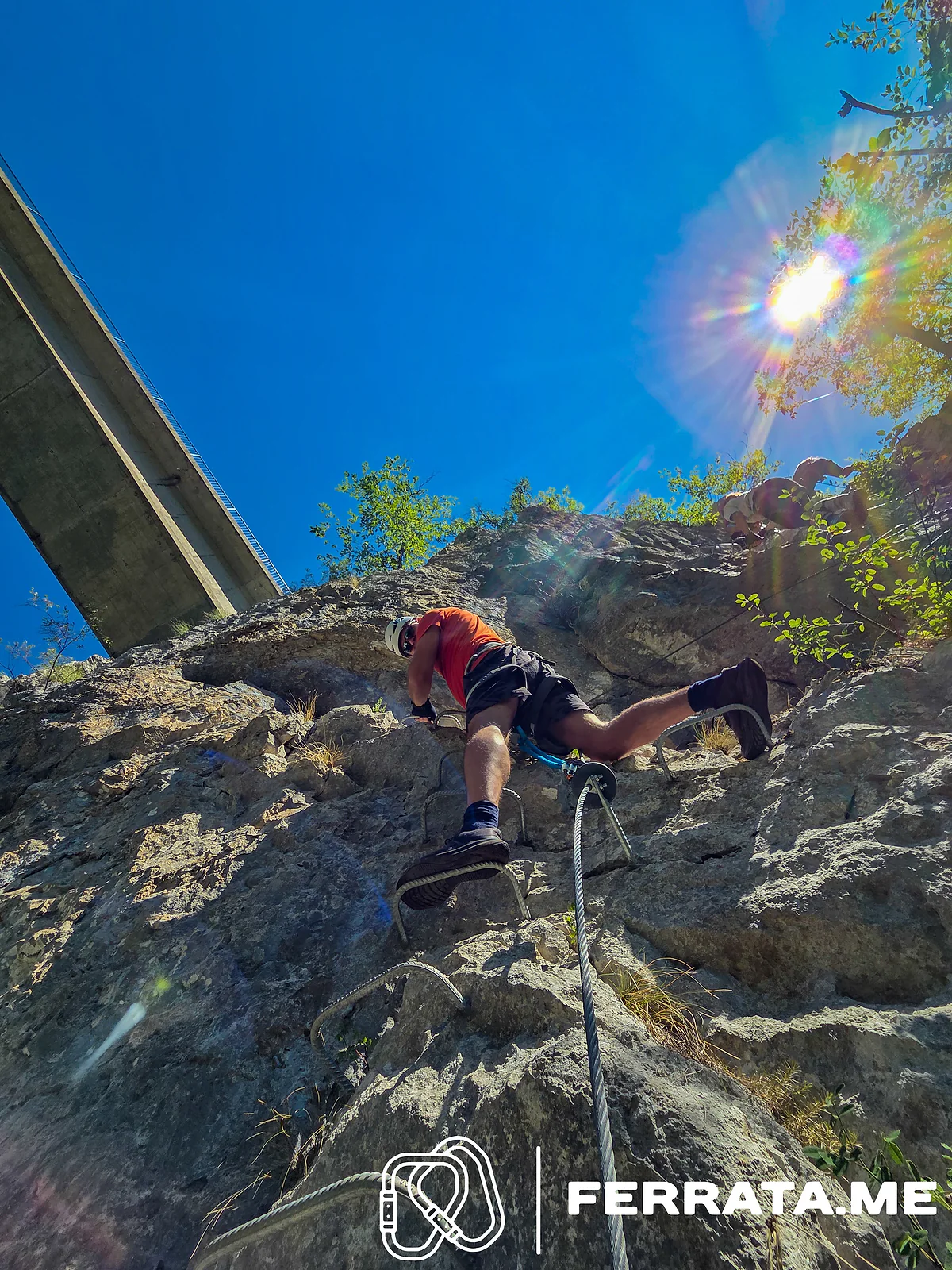 A climber in a white helmet and orange T-shirt pulls up over a bulge on the iron rungs, clipped to the cable, with the concrete bridge high overhead against a blue sky and the sun behind a tree.