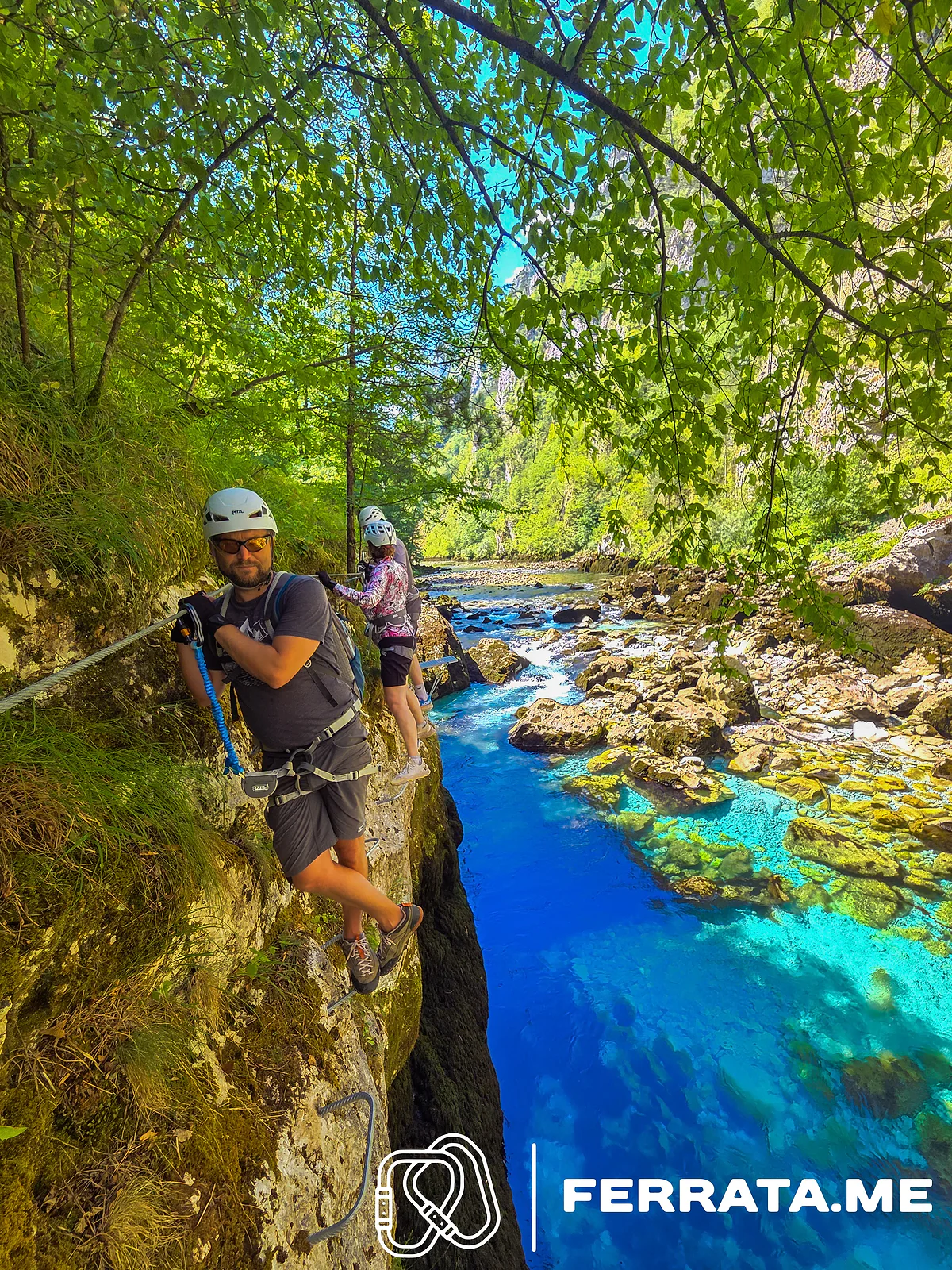 A man in a white helmet holds the blue arm of his via ferrata set clipped to the cable on a mossy wall above a deep blue stretch of the Piva, with two climbers in white helmets behind him.