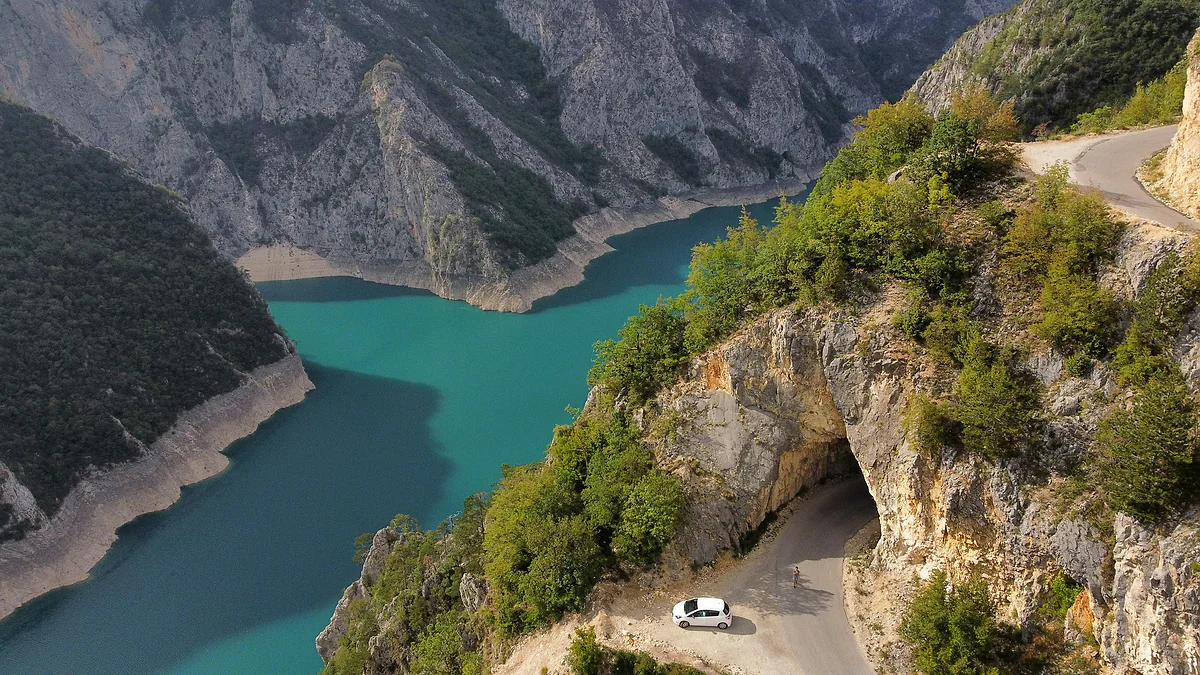 A white car parked on a narrow road at the mouth of a tunnel cut into a limestone cliff, high above the turquoise water of the reservoir, with grey rock walls on the far shore.