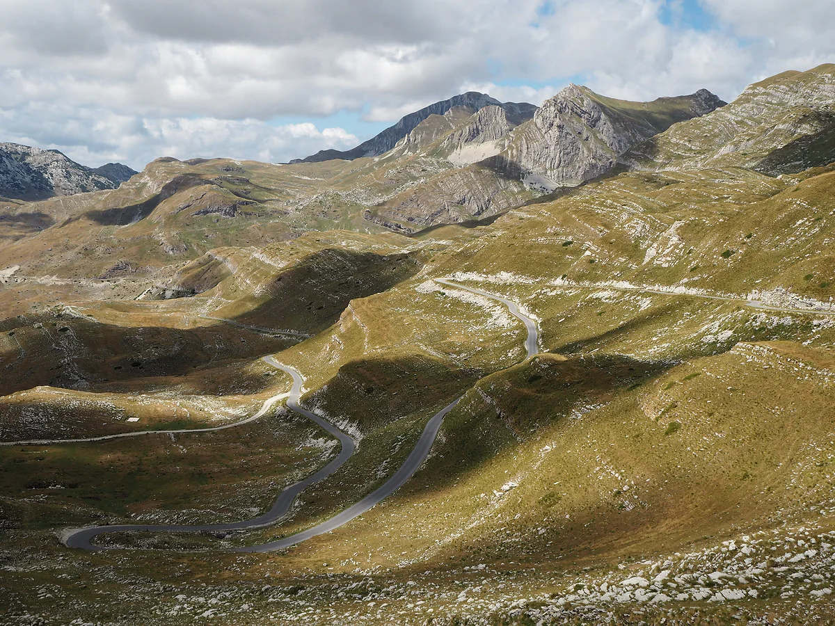 A paved mountain road winding in loops across brown summer grassland on the Durmitor plateau, with grey limestone peaks behind under a cloudy sky.