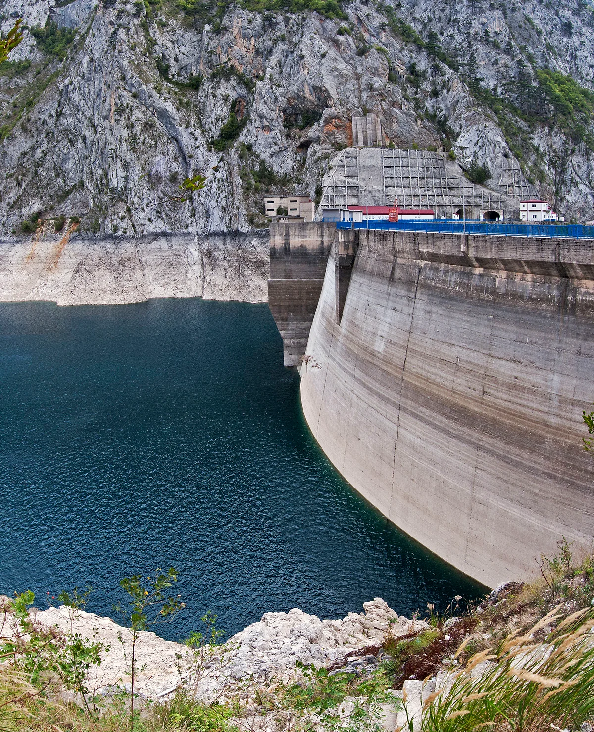 The curved grey concrete wall of the Mratinje Dam rising out of dark blue water, with the road and blue railings along its crest and a limestone cliff behind.
