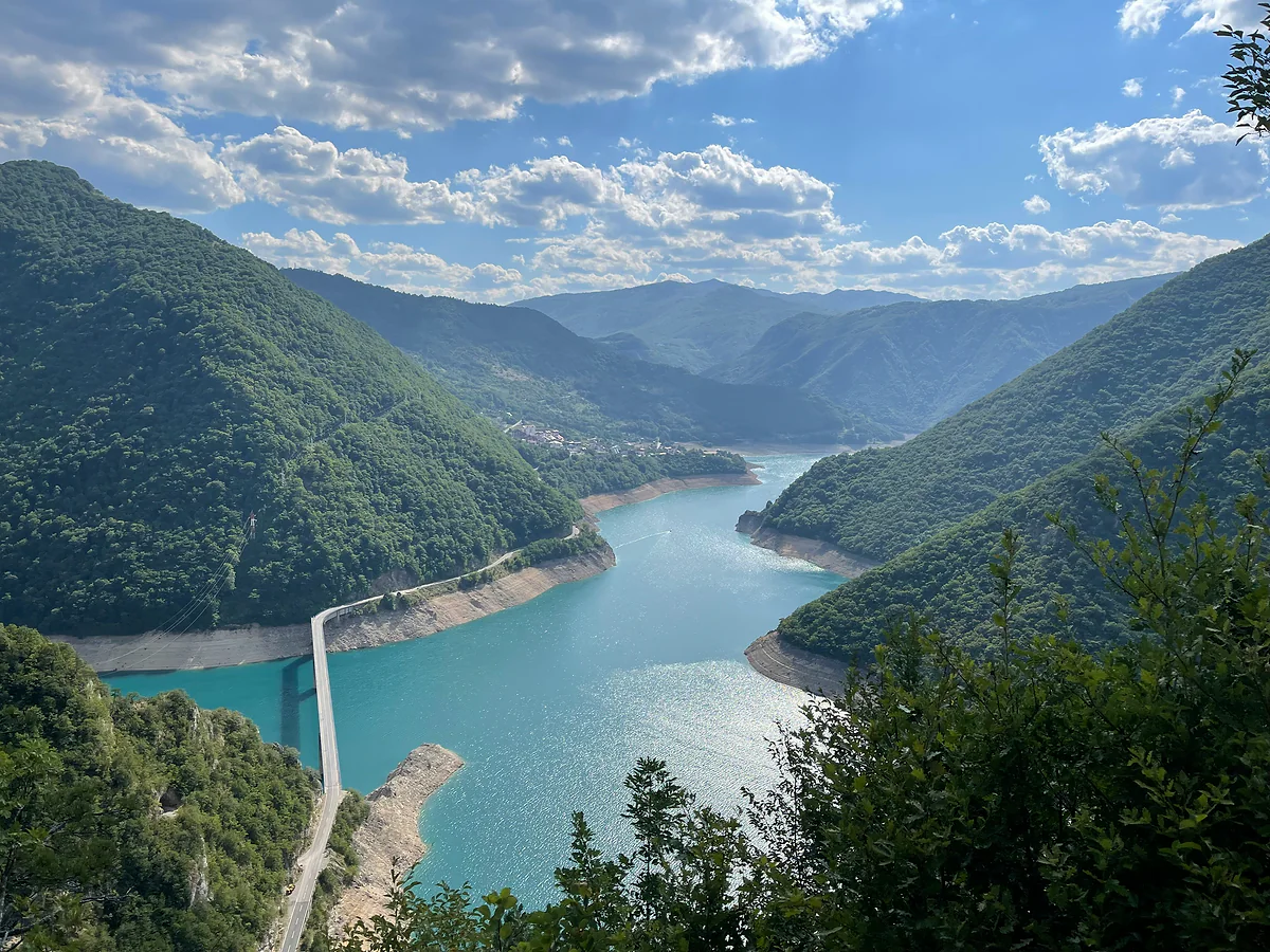 Piva Lake from a wooded slope above Plužine, with a long road bridge crossing the turquoise water and forested mountains on both sides under white clouds.