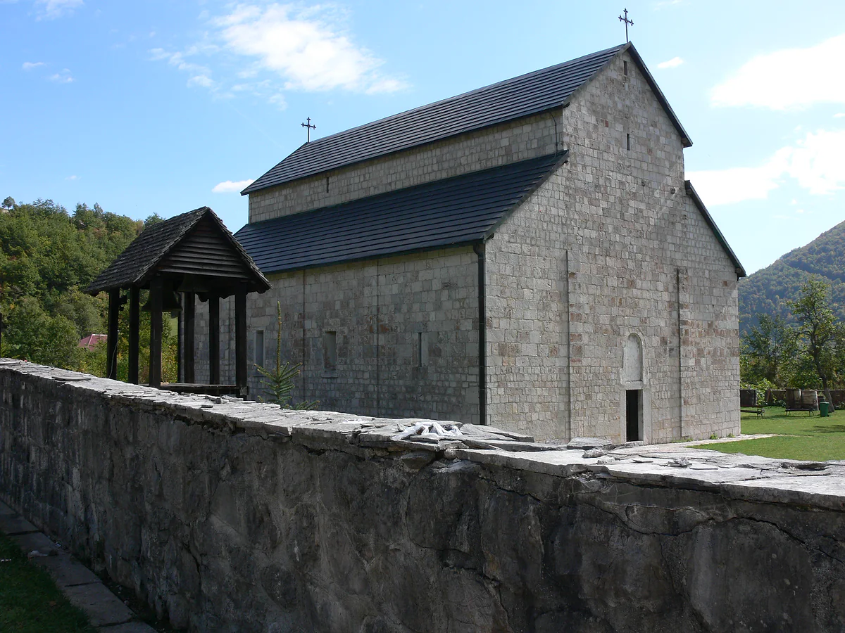 The grey stone church of Piva Monastery with a dark tiled roof and crosses on top, behind a low stone wall, with a small roofed wooden structure beside it and green hills under a blue sky.