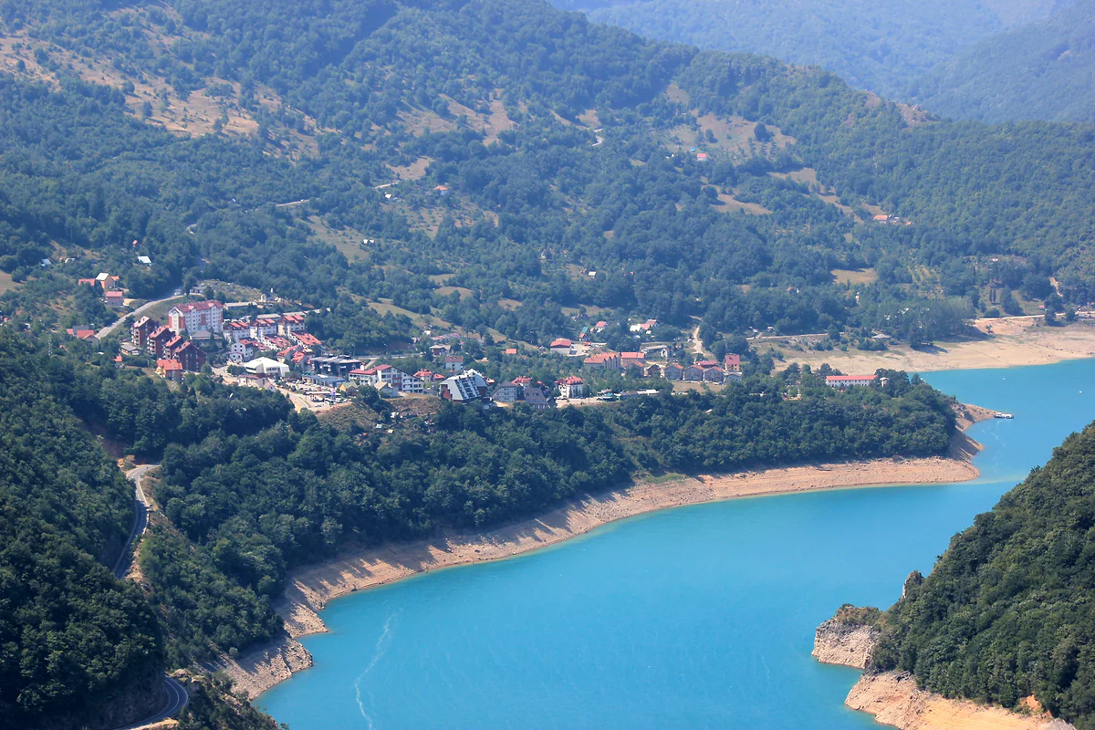 Plužine seen from above, a cluster of houses and apartment blocks with red roofs on a forested hillside over the turquoise lake, with a band of pale rock along the shore.