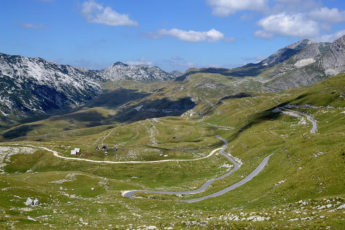 A narrow paved road winds in hairpins across green grassland on the Durmitor plateau, past a few wooden huts, with grey limestone peaks behind under a blue sky.
