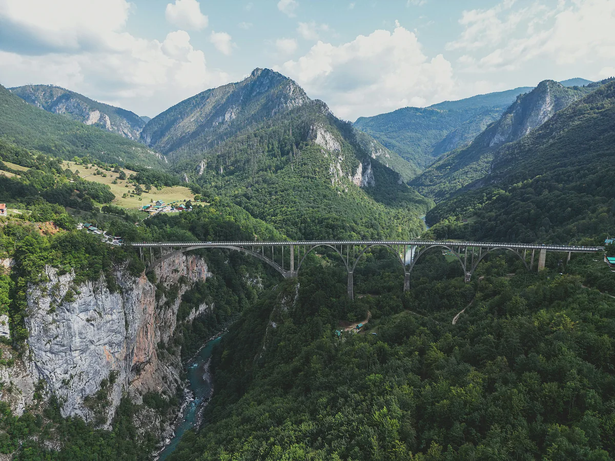 A long concrete bridge on five arches spans a deep forested canyon, with the Tara a thin turquoise line far below and mountains behind under a cloudy sky.