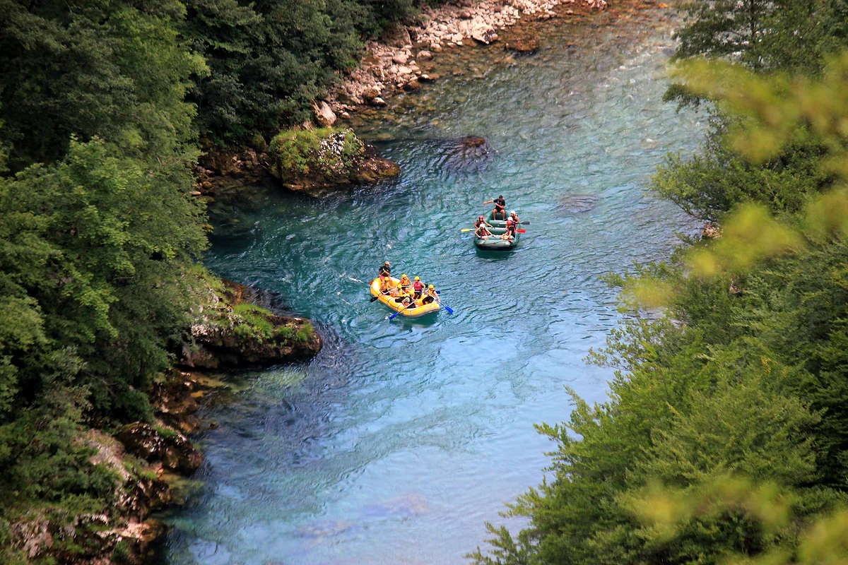 Two rafts with paddlers in yellow helmets float on clear turquoise water between forested banks, seen from high above.