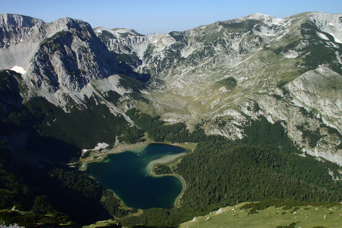 A dark blue lake in the shape of a heart lies in a forested hollow, seen from high on a grassy slope, with bare grey limestone mountains rising all round it.